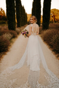 mariée dans sa magnifique robe blanche avec une longue traîne brodée. elle est de dos avec son bouquet de fleurs dans la main, elle est au milieu d'une allée de grands cyprès au mas des costes en provence