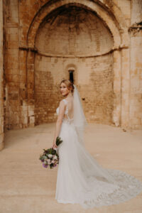 mariée dans sa robe de mariée blanche avec des broderies et dentelles avec son très long voile et son bouquet de fleurs à la main